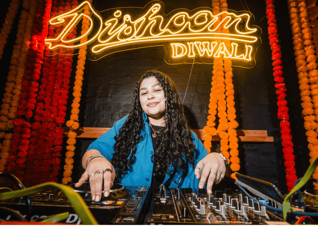 A DJ plays tunes on decks with Dishoom Diwali lit up in the background